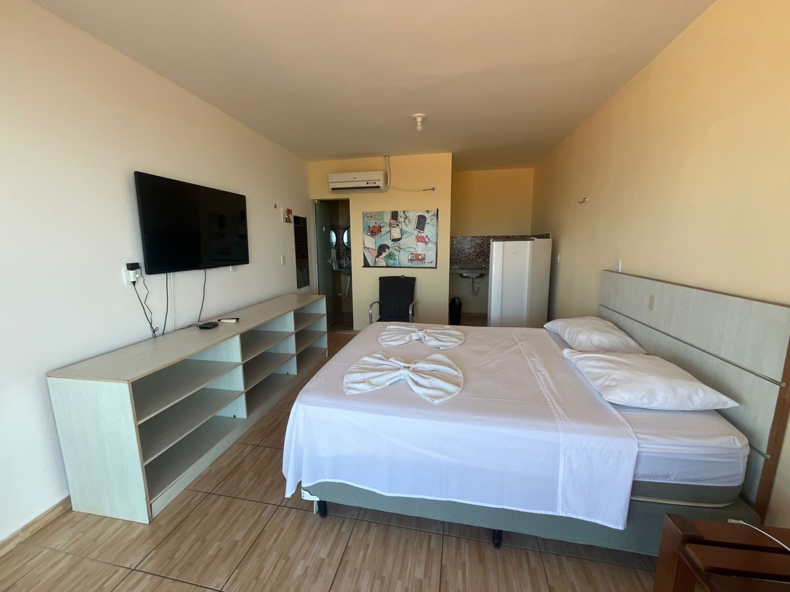 A hotel room with a bed, a TV mounted on the wall, a small desk, and a kitchenette in the background.