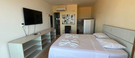 A hotel room with a bed, a TV mounted on the wall, a small desk, and a kitchenette in the background.