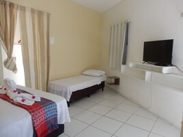 Una habitación de hotel con cama, televisor en un estante, mesita de noche pequeña y ventana con cortinas.