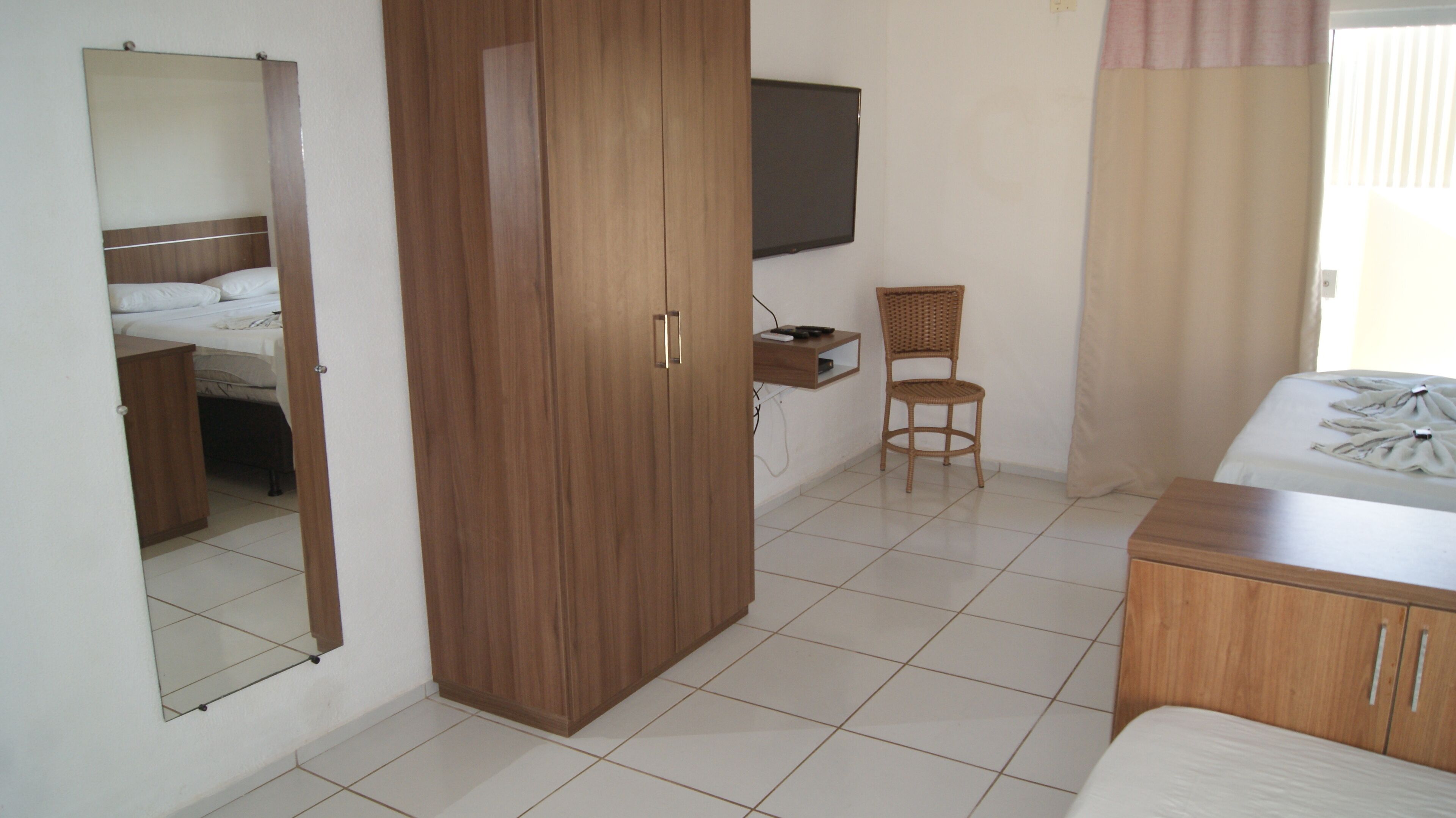 Habitación de hotel con cama, armario de madera, televisor montado en la pared, una mesita pequeña, una silla de mimbre y una ventana con cortinas.