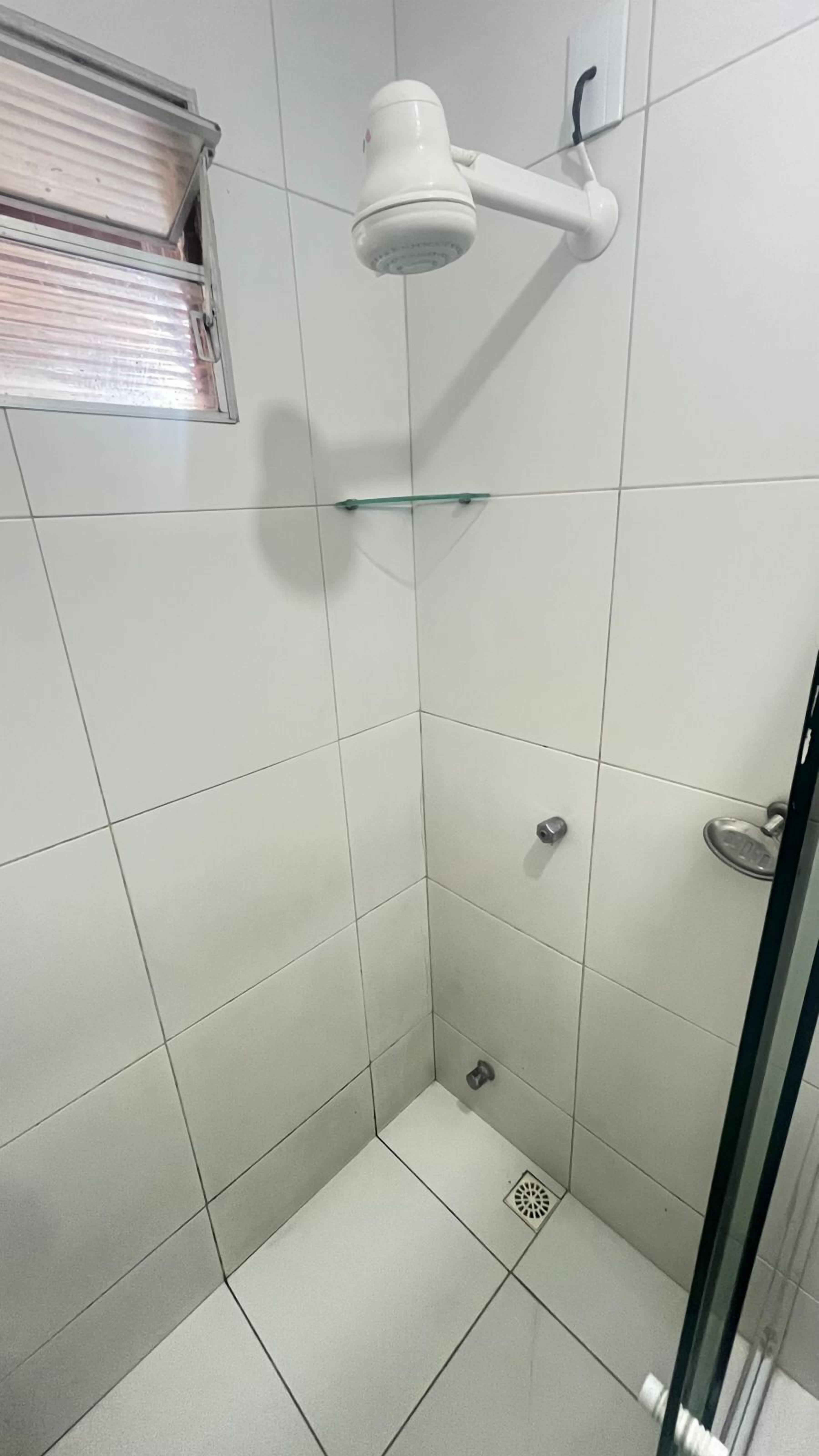 Pequeno espaço de duche revestido com azulejos, com chuveiro manual fixado na parede, prateleira de vidro e ralo.