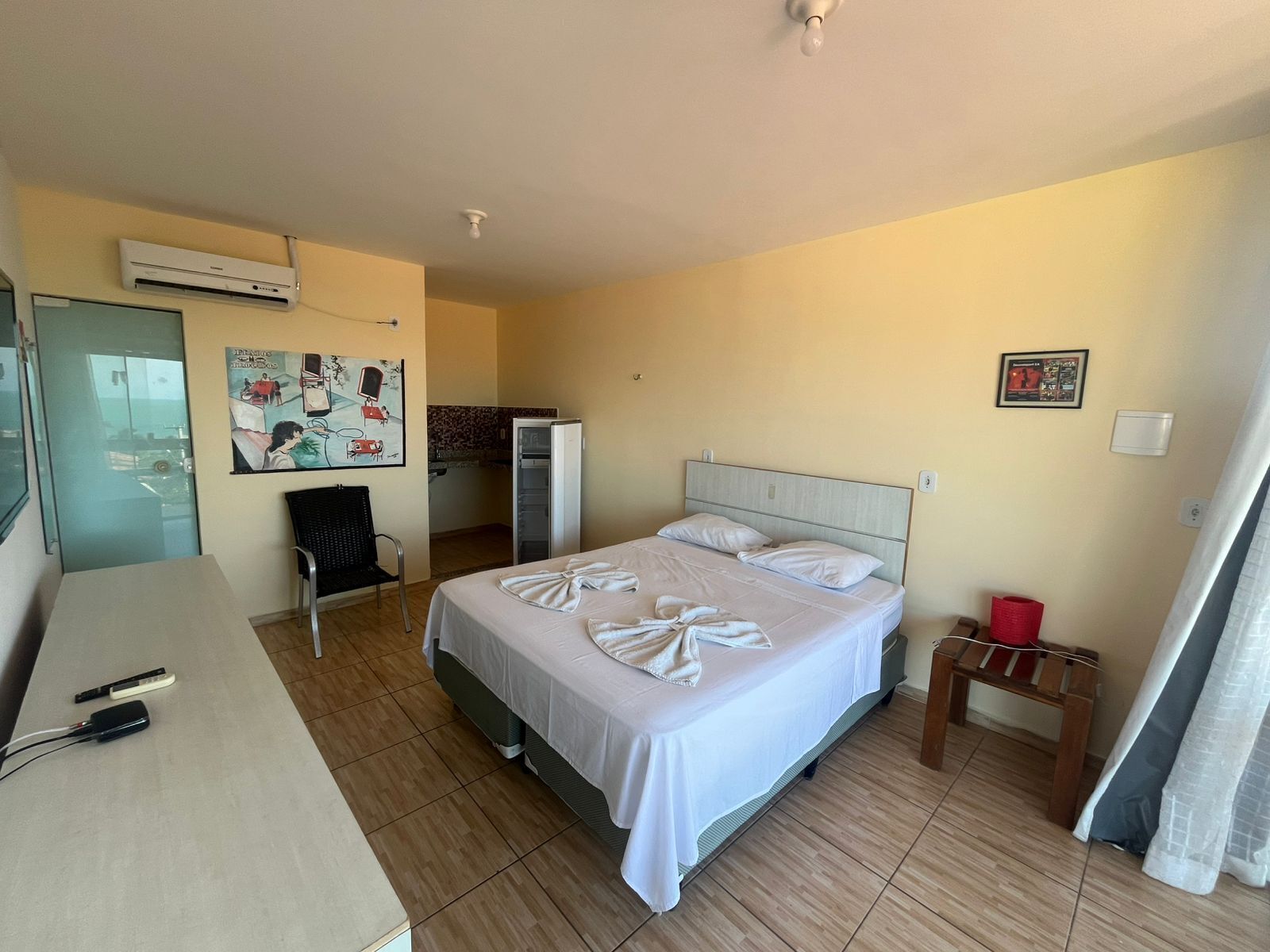 A hotel room with a bed, a desk, an air conditioner, a chair, a refrigerator, and a picture on the wall.