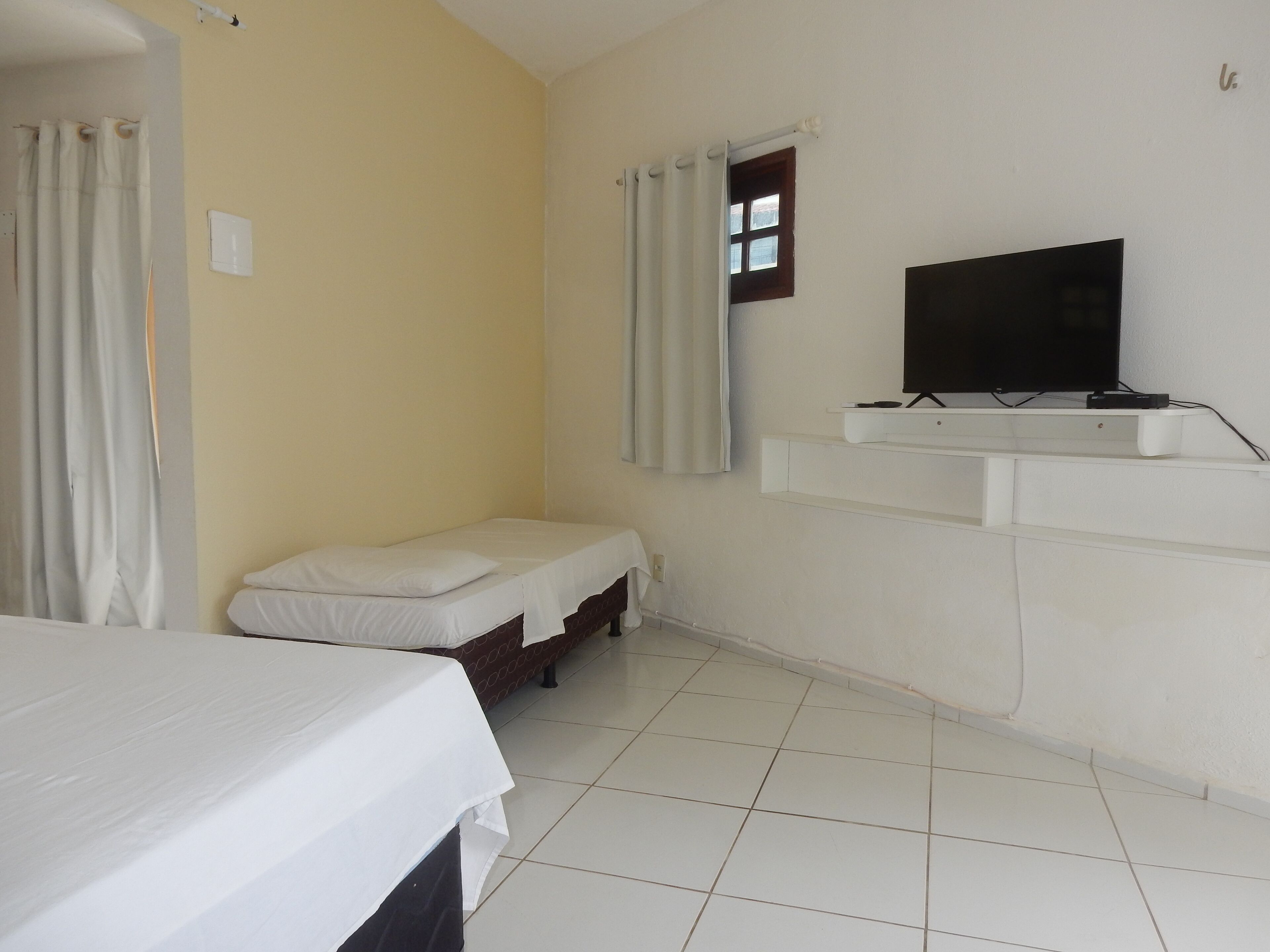A small, clean room with a single bed, a television mounted on the wall, and a window with curtains.