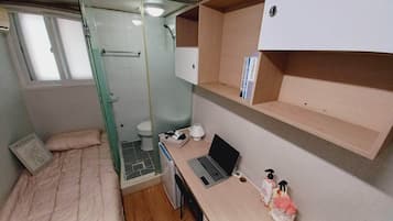 A compact room with a bed, a shower, a toilet, a desk with a laptop, and shelves with books.
