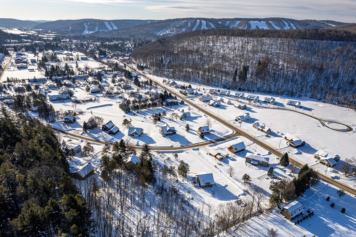 An aerial view of a snow-covered residential area with houses, roads, and a forested mountain backdrop.