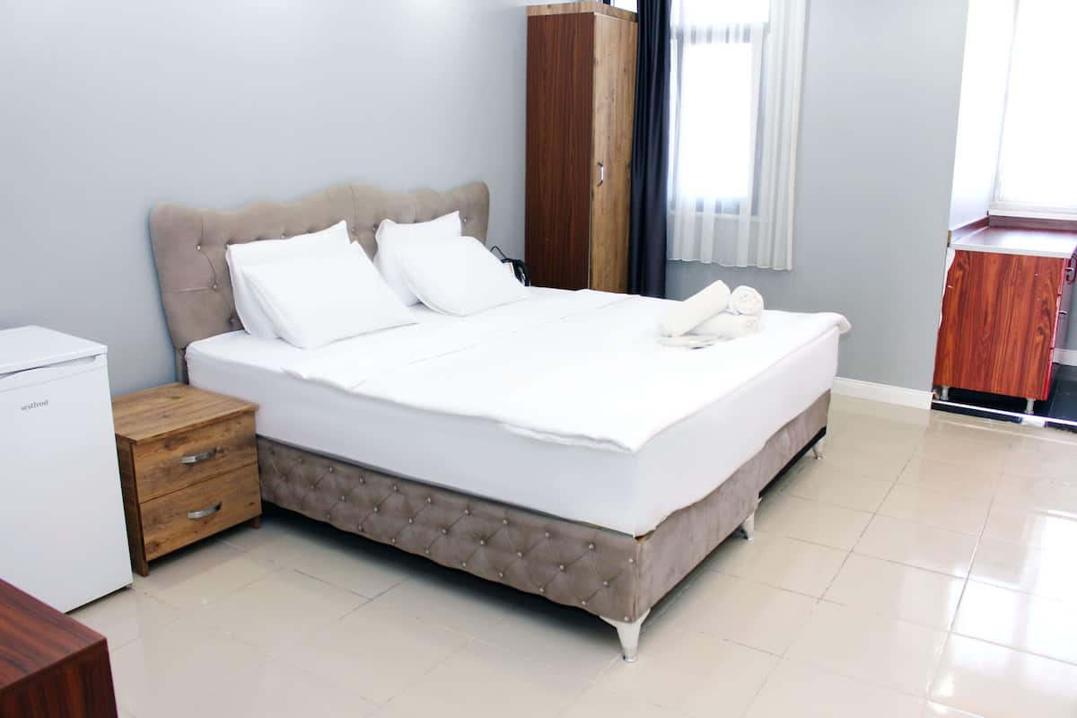 A neatly made bed with white linens, a wooden nightstand with drawers, a wardrobe, and a small white appliance.