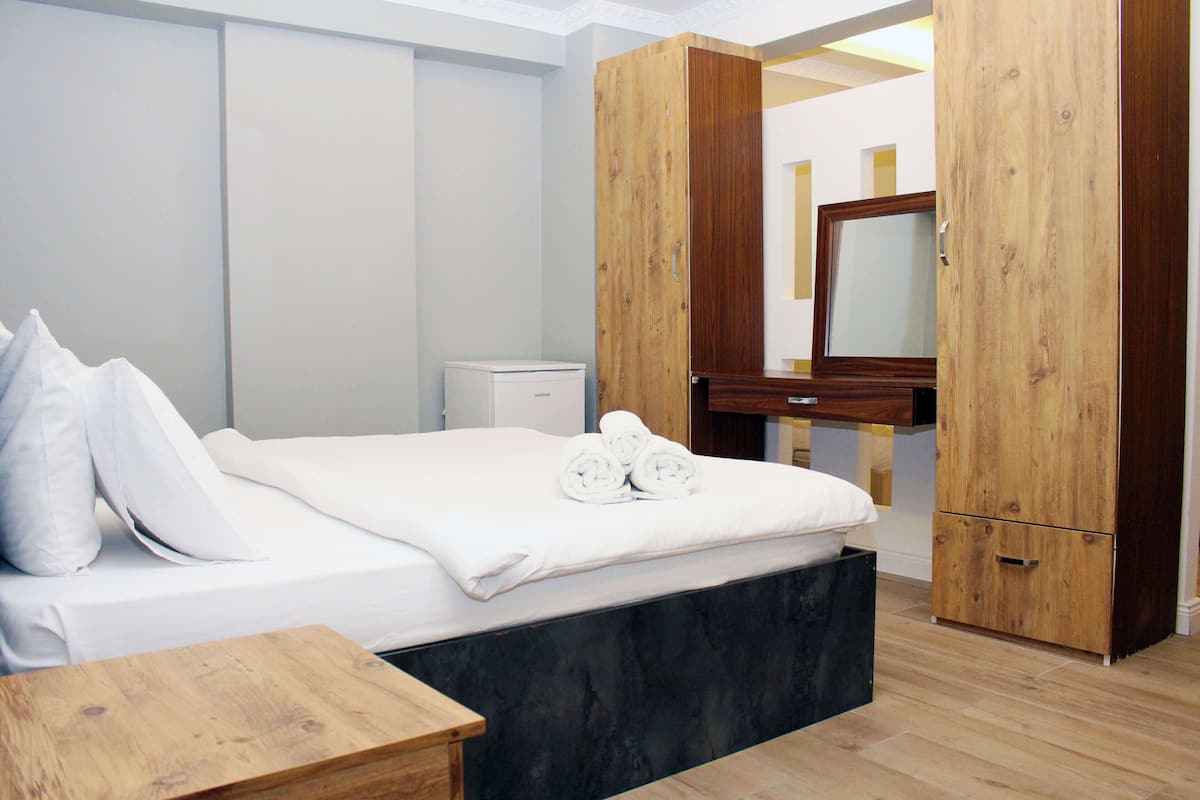 A bedroom with a bed, a wooden nightstand, a wooden wardrobe, a small bedside table, and a mirror.