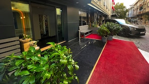 Entrada de hotel con alfombra roja, plantas en macetas y un banco.