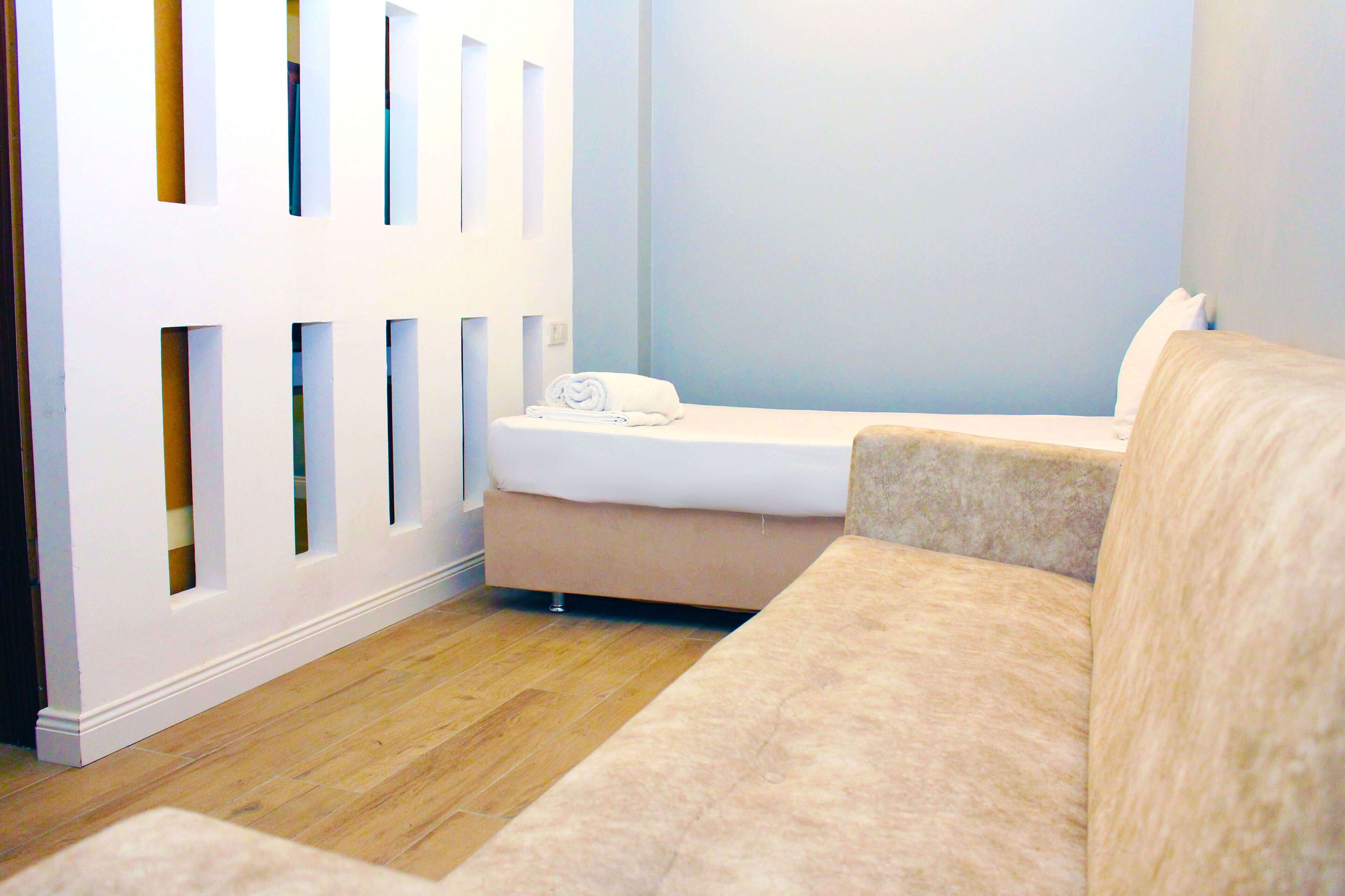 Una stanza con un divano beige, un letto piccolo con materasso bianco e un asciugamano bianco arrotolato, e un pavimento in legno.