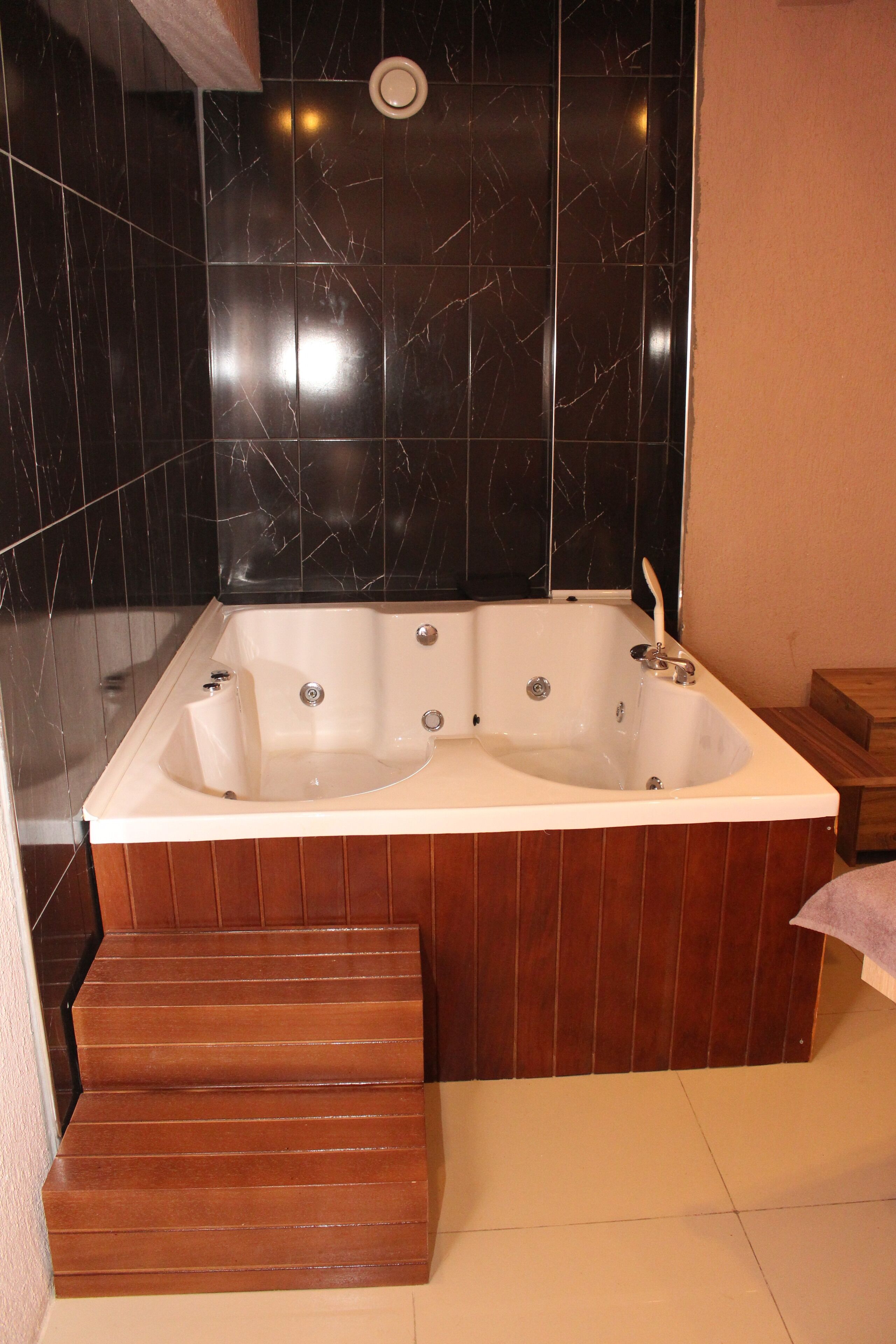 Un baño con una bañera grande, escalones de madera que la conducen y paredes de mármol negro.