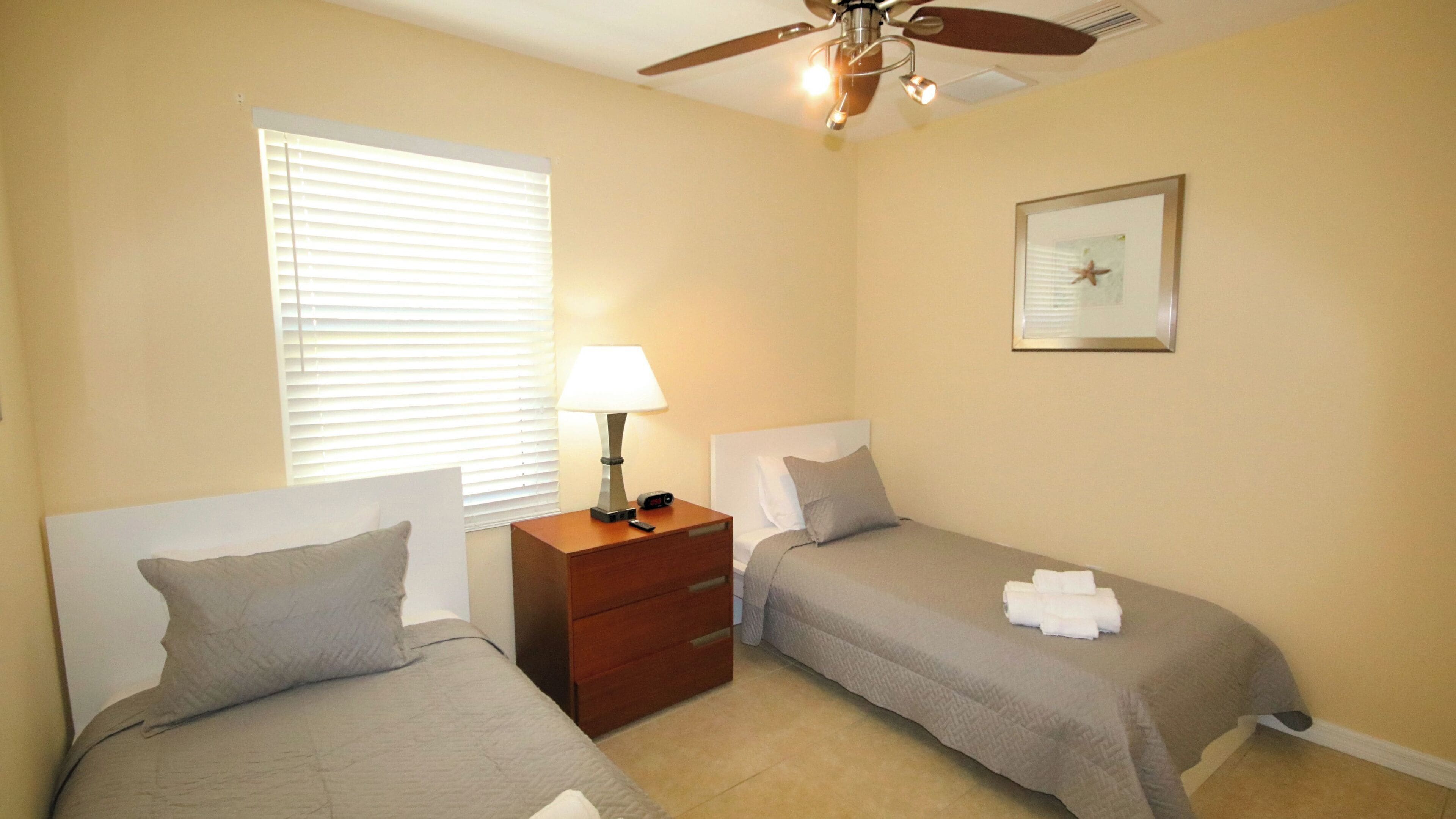 A hotel room with two beds, a nightstand with a lamp, a ceiling fan, and a framed picture on the wall.