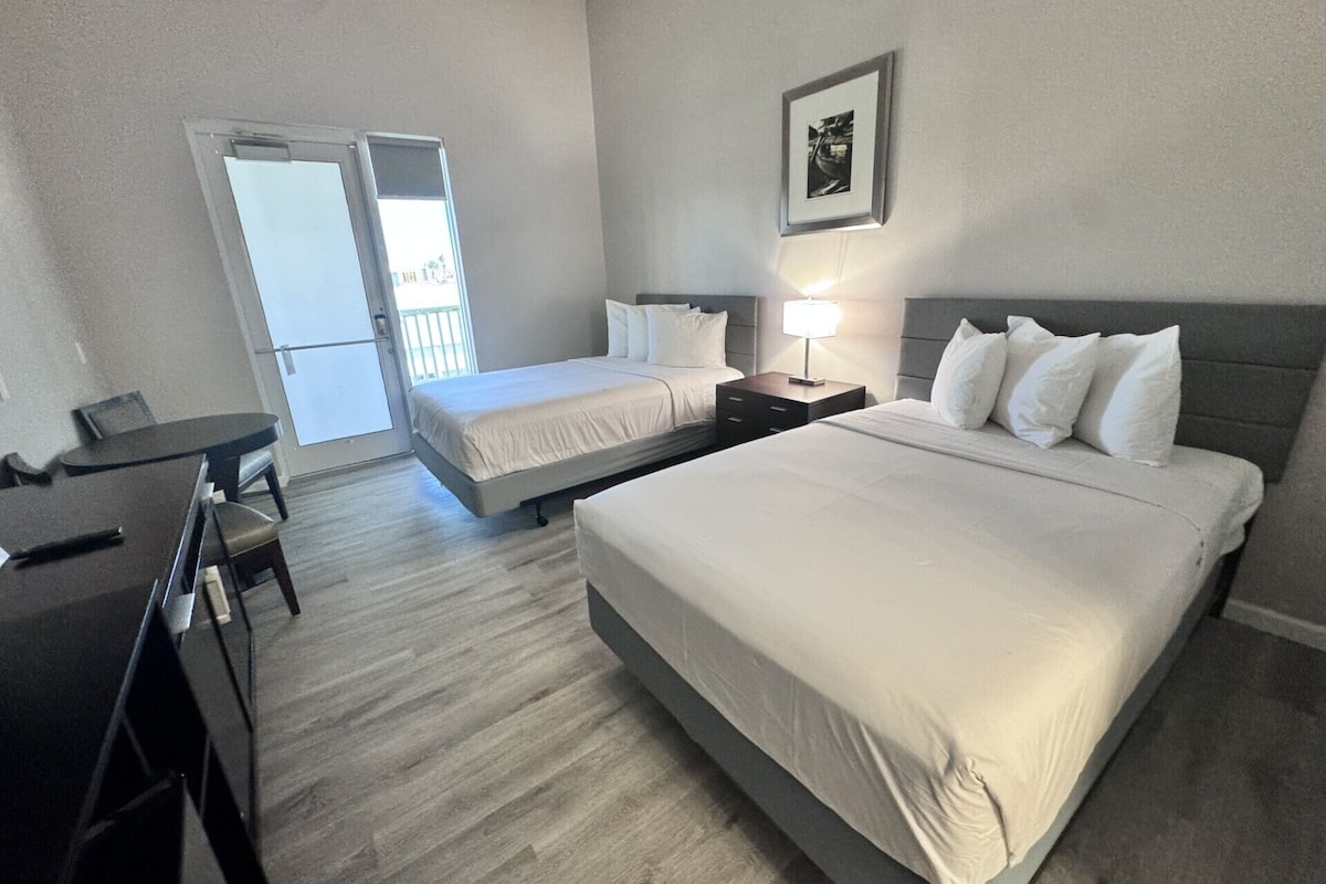 Habitación de hotel con dos camas, un escritorio y una silla. Hay una puerta que da a un balcón.