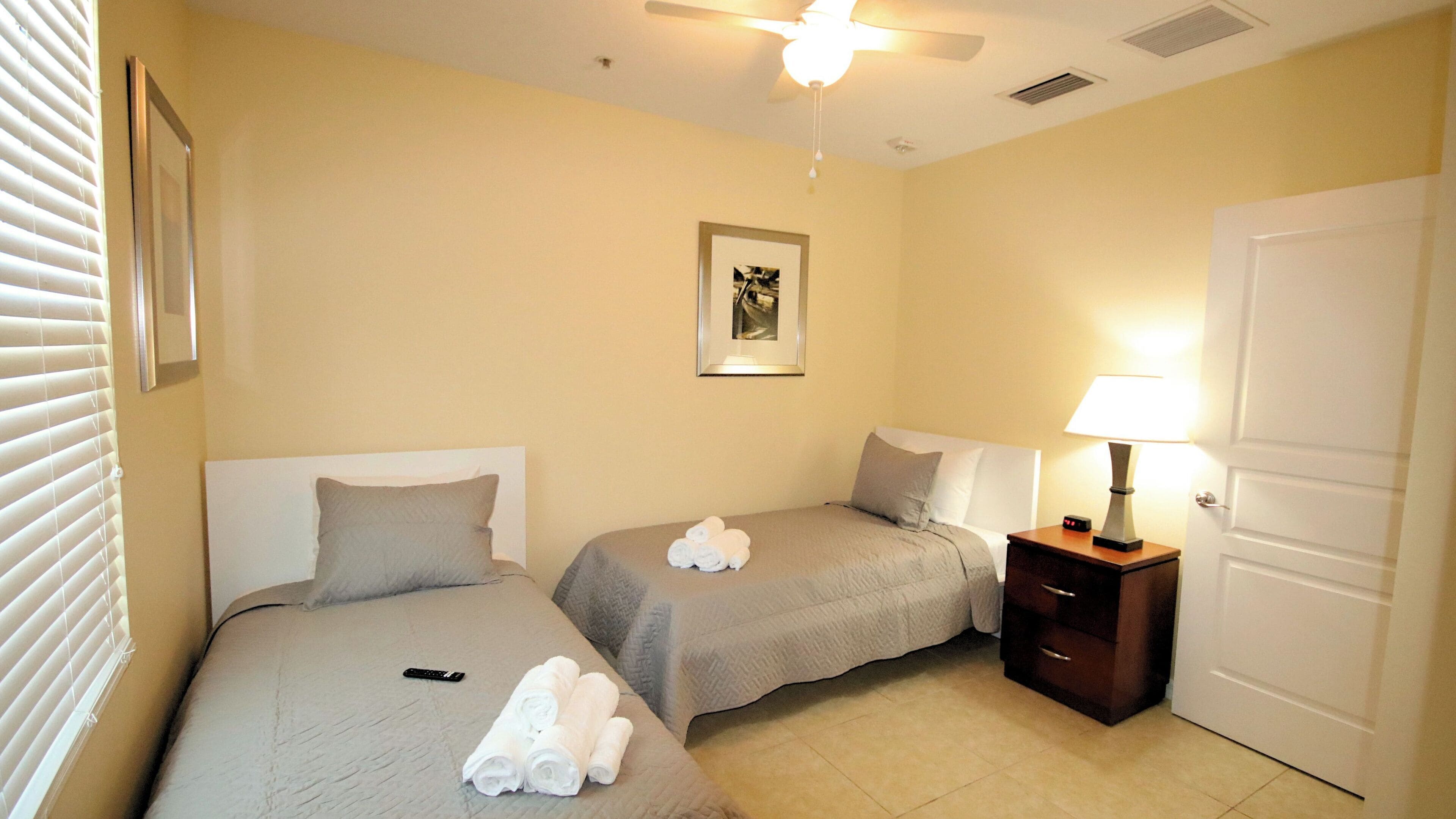 A hotel room with two beds, a ceiling fan, a framed picture on the wall, a bedside table with a lamp, and a door.