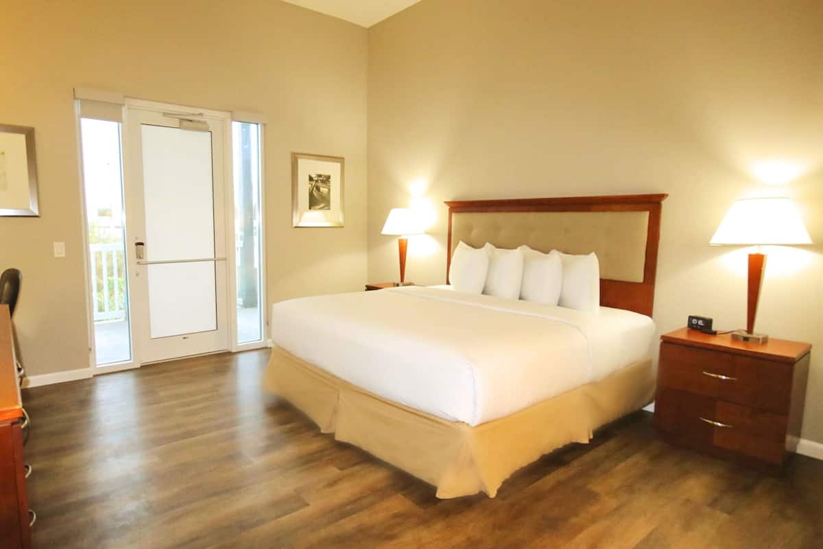 Habitación de hotel con una cama grande, dos mesitas de noche con lámparas, un escritorio con una silla y una puerta que da a un balcón.