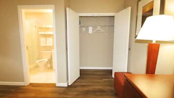 A hotel room with a wooden floor, a bathroom with a toilet and sink, a closet with a hanging rack, and a bedside table with a lamp.