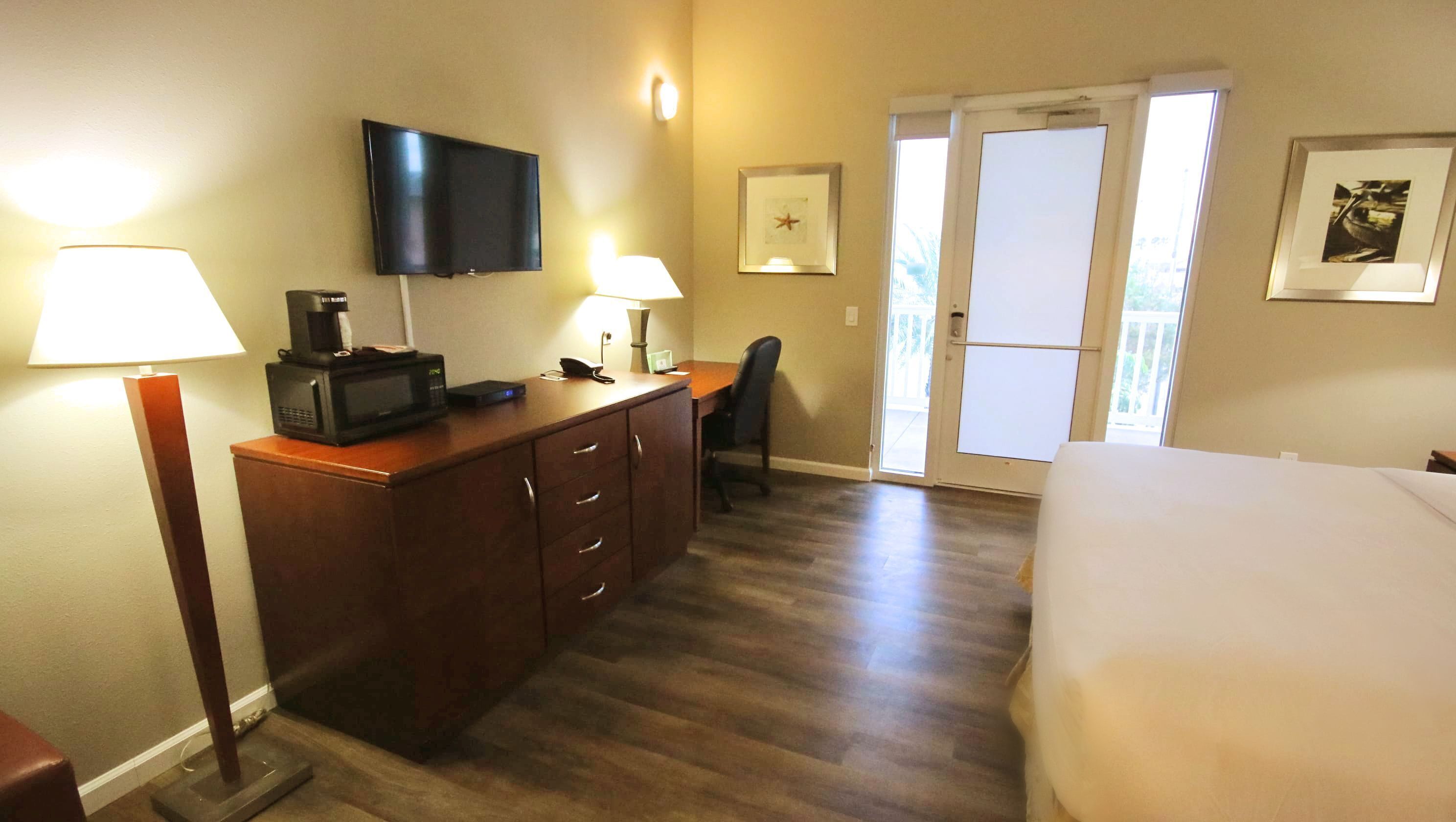 A hotel room with a bed, a desk with a flat-screen TV, a microwave, and a coffee maker. There is a chair, a door leading to a balcony, and a framed picture on the wall.
