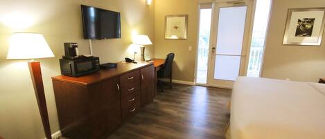 A hotel room with a bed, a desk with a flat-screen TV, a microwave, and a coffee maker. There is a chair, a door leading to a balcony, and a framed picture on the wall.