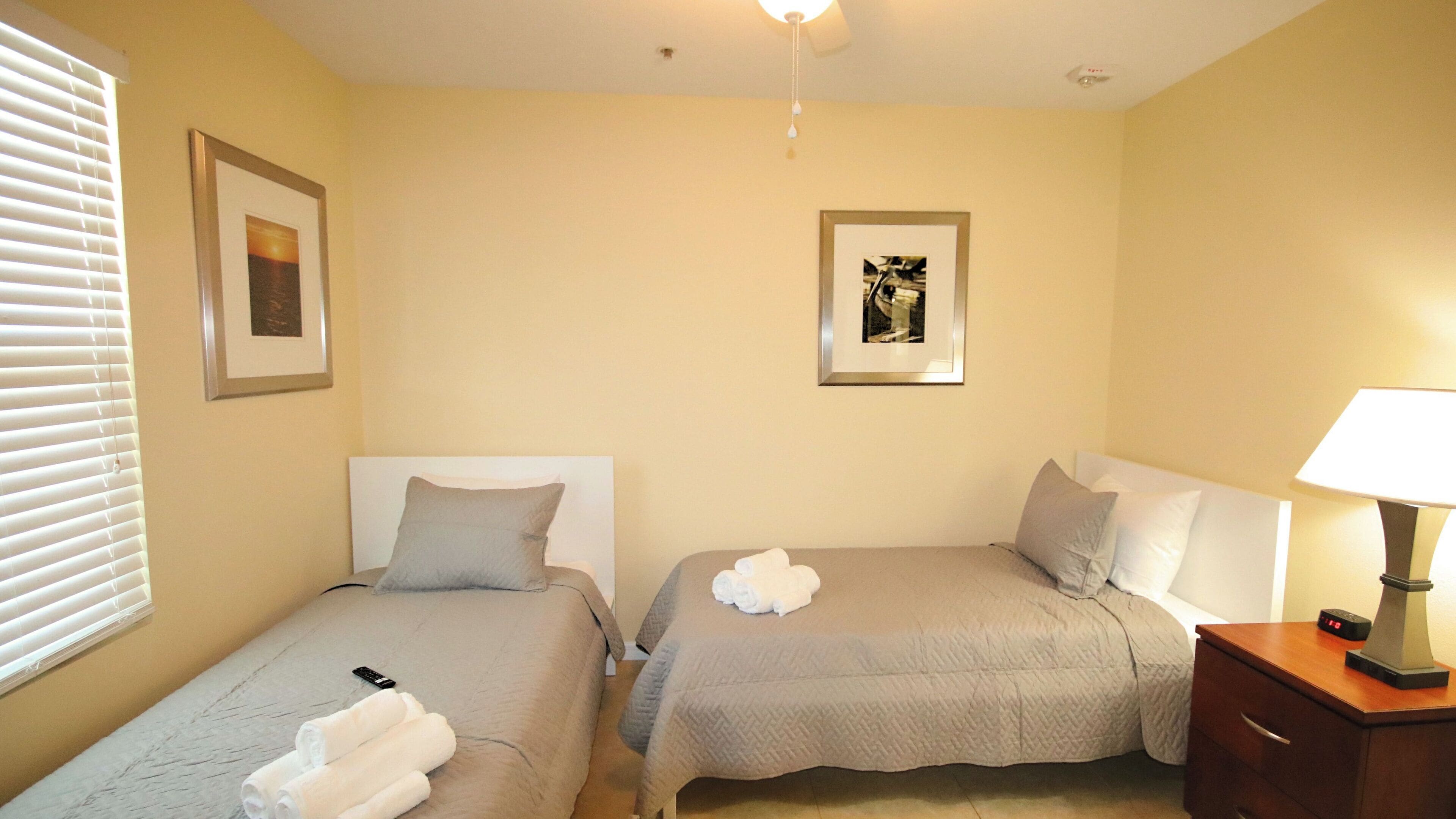 A hotel room with two single beds, a bedside table with a lamp, a clock, and two framed pictures on the wall.