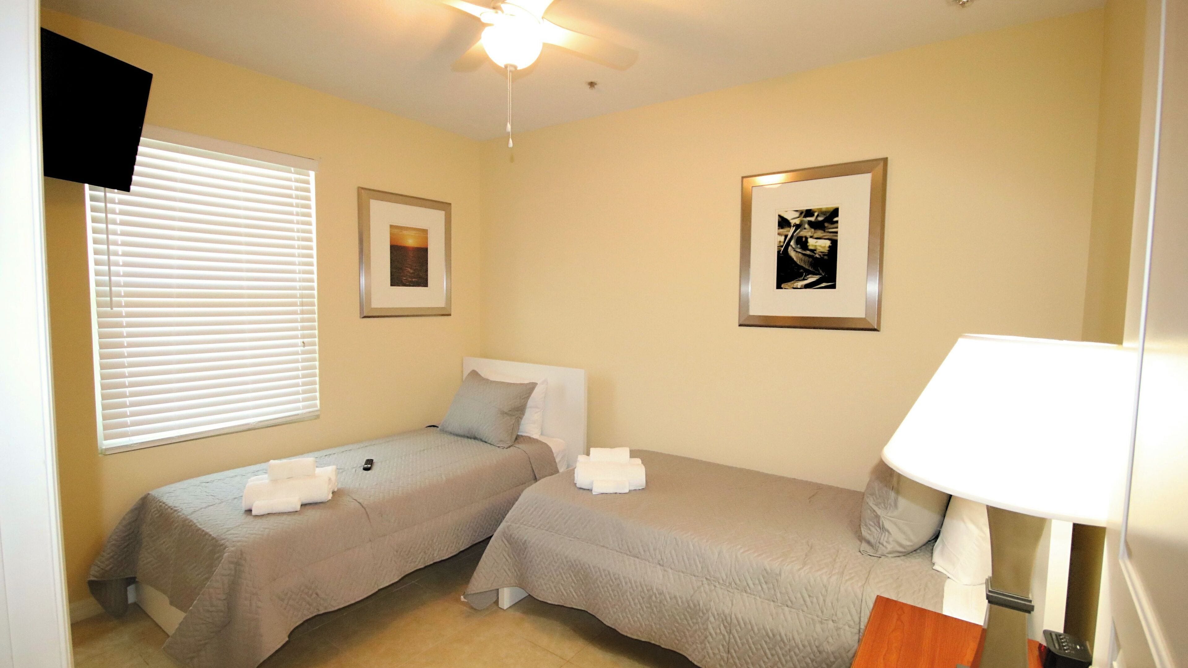 A bedroom with two single beds, a ceiling fan, a window with blinds, and two framed pictures on the walls.