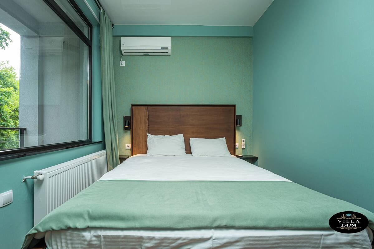Un dormitorio con cama, ventana, aire acondicionado y vistas a la vegetación.
