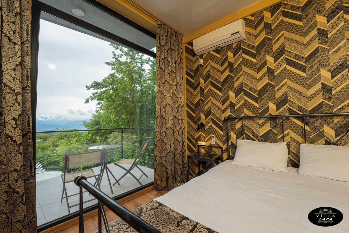Habitación de hotel con una ventana grande, una cama, una mesita de noche y vistas a zonas verdes y montañas.