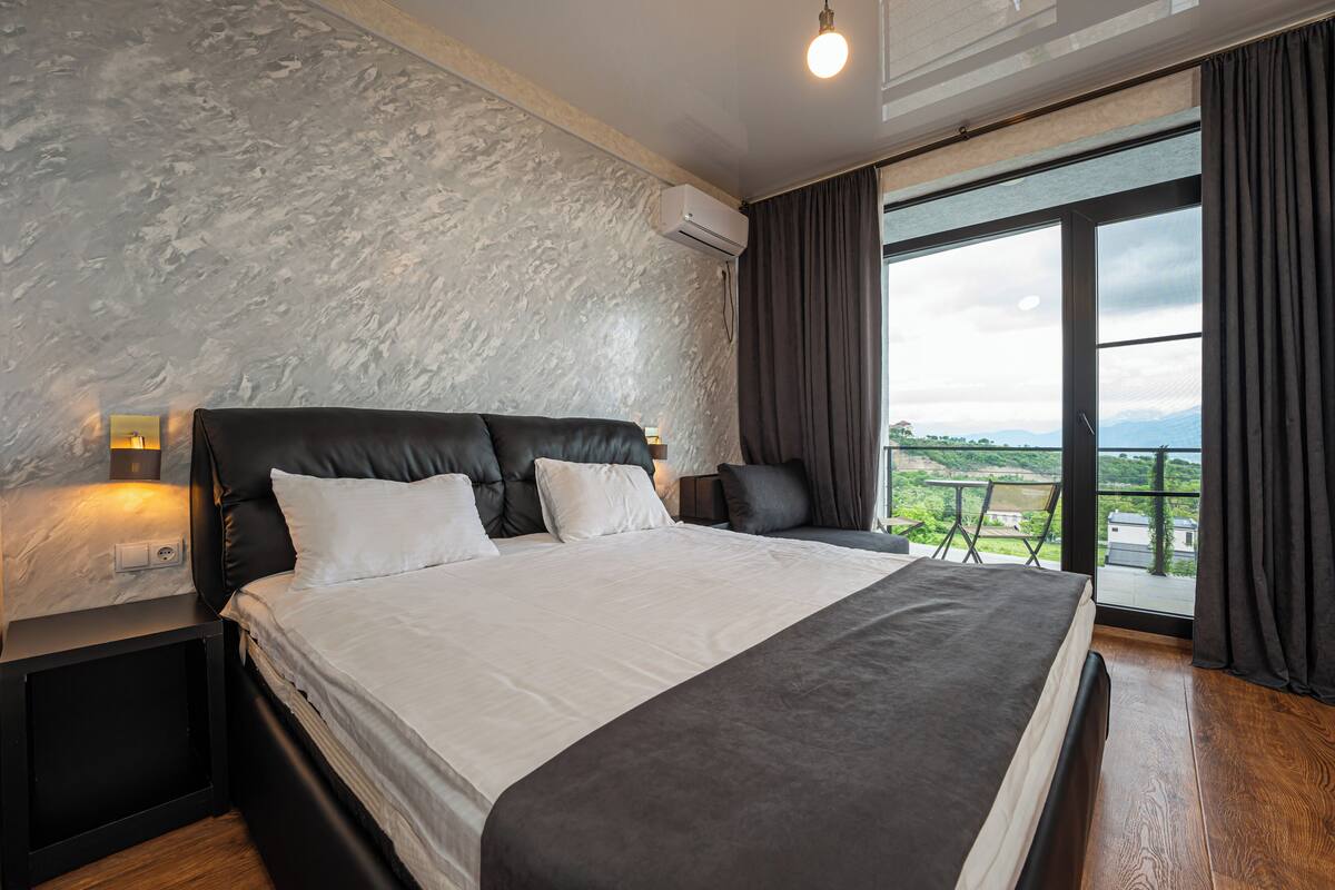 Un dormitorio moderno con una cama grande, cabecera, mesita de noche y vistas al exterior a través de un ventanal amplio.