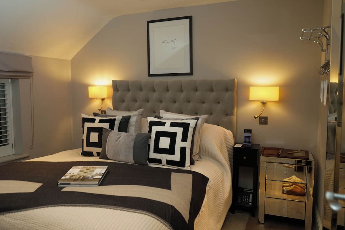 A neatly arranged bedroom with a tufted headboard, two bedside lamps, a framed picture on the wall, and a bedside table with a lamp and a book.