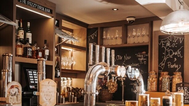 Uma área de bar com um letreiro que diz "The Fuzzy Duck Bar", uma torneira de cerveja rotulada como "Gulf" e um cardápio em lousa com itens como "Our Nachos" e "Fuzzy Duck Nachos".