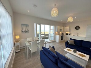A modern living room with a blue sofa, a dining area with white chairs, and a kitchen in the background.