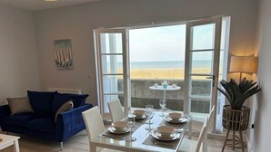 Uma sala de jantar com mesa posta para quatro pessoas, cadeiras brancas e vista para a praia através de grandes janelas.