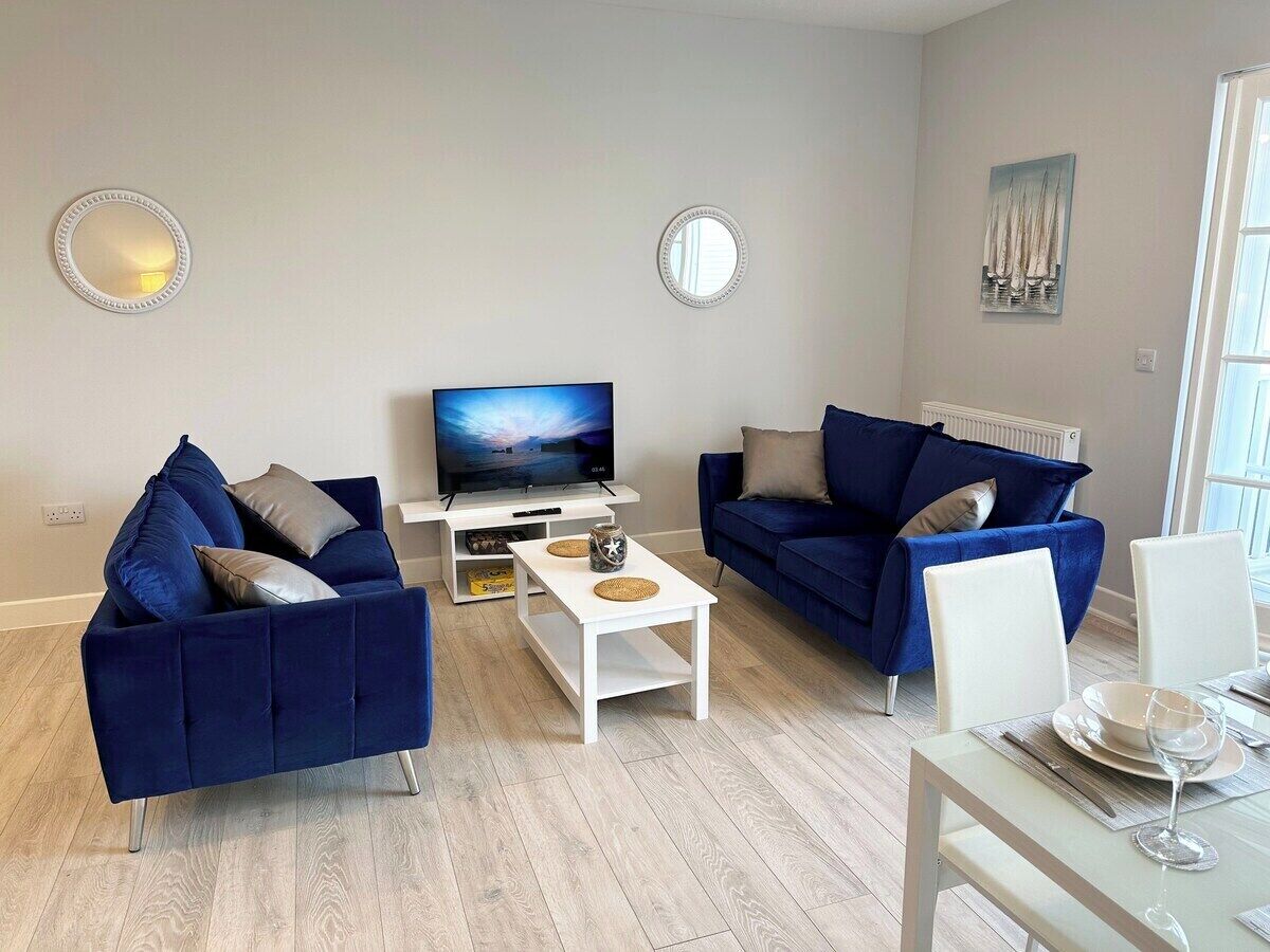 A modern living room with two blue sofas, a white coffee table, a TV, and a dining area with a table set for two.