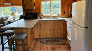 A kitchen with wooden cabinets, a microwave, a refrigerator, a sink, and a window with a view of trees. - Cozy Cabin on Rangeley Lake  (Rangeley)