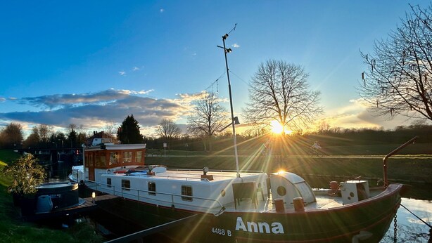 A boat named 'Anna' docked near a grassy area with a clear sky and sun setting in the background. - Au fil de Leau (Plobsheim)