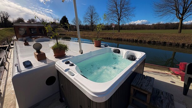 A hot tub with a wooden exterior, a small deck area with potted plants, and a view of a canal with trees and a building in the background. - Au fil de Leau (Plobsheim)