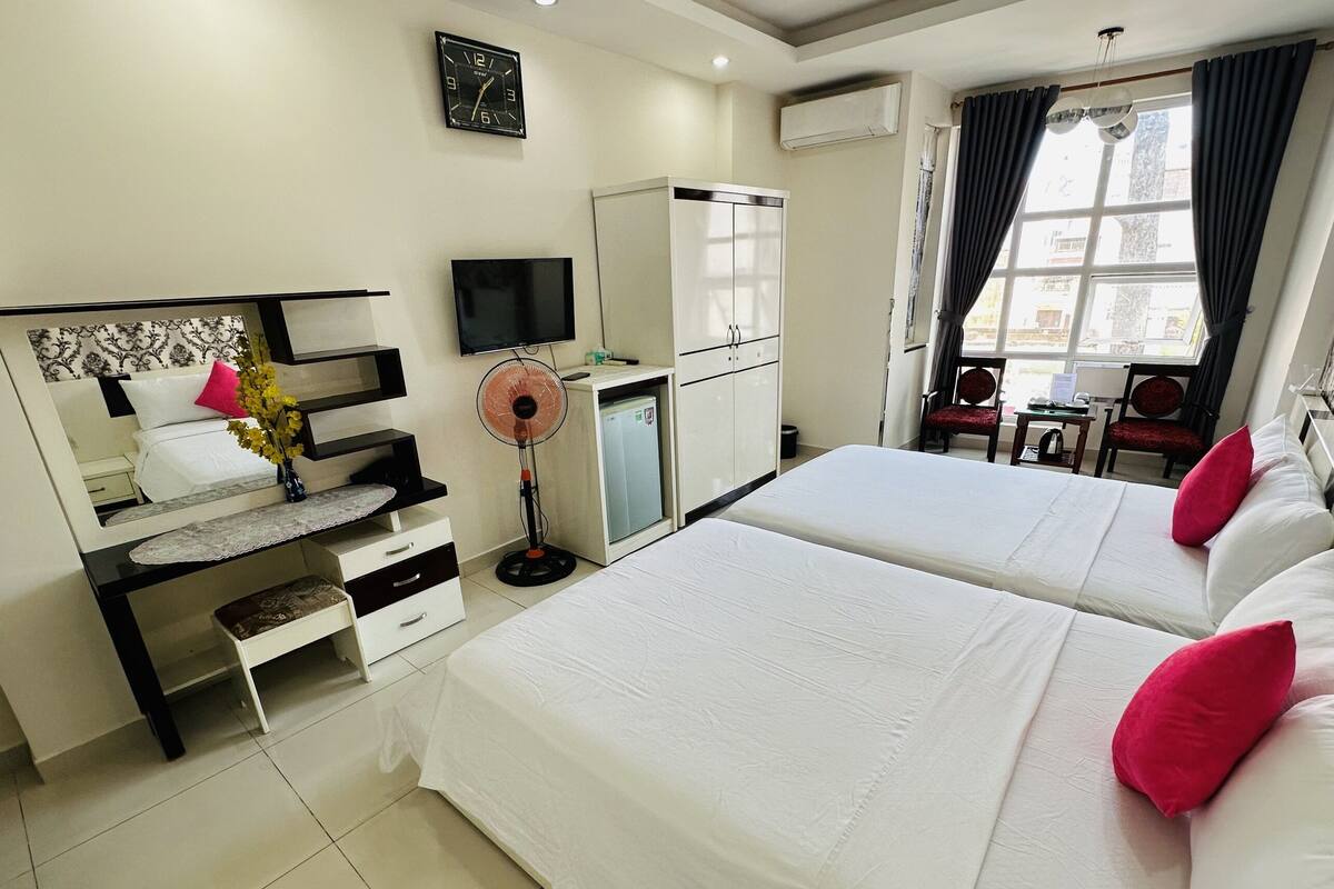 Habitación de hotel con dos camas, un escritorio con silla, televisor, armario y ventana con cortinas.