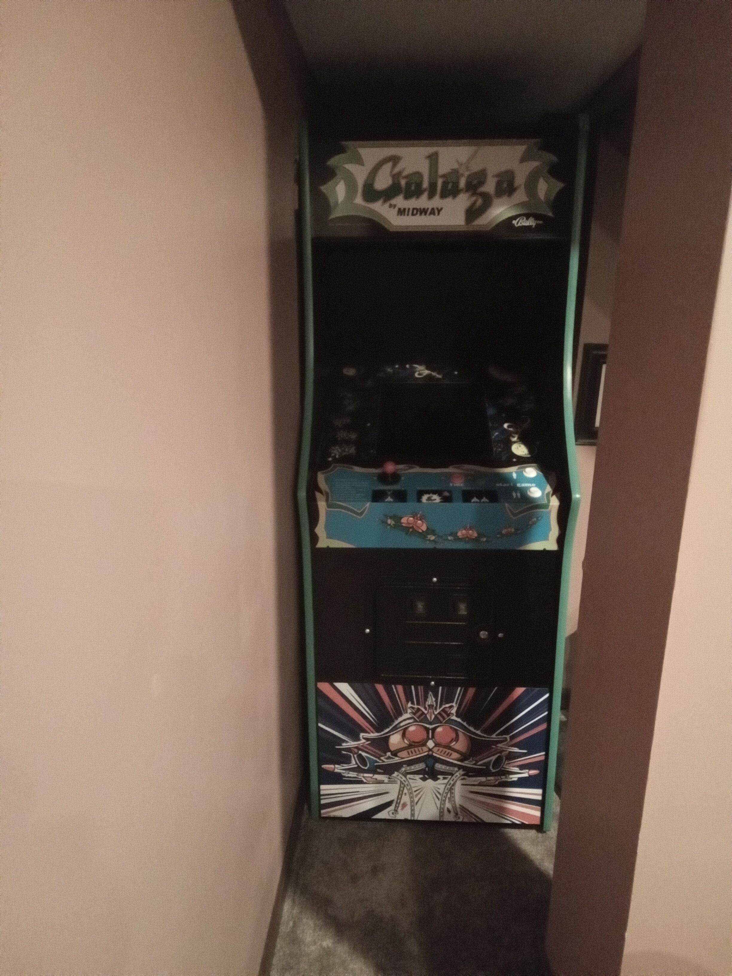Un pasillo estrecho con una máquina de videojuegos antigua, identificada como "Galaga" de Midway.
