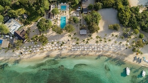 An aerial view of a resort with a large pool, beach area with lounge chairs, and palm trees.