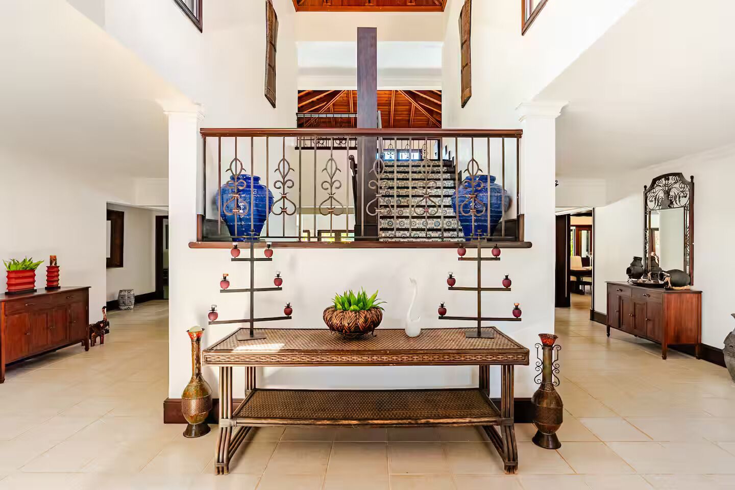 Un hall d’entrée spacieux, doté d’un escalier central, d’une rampe décorative en métal et de divers meubles, notamment une table en bois et un buffet.