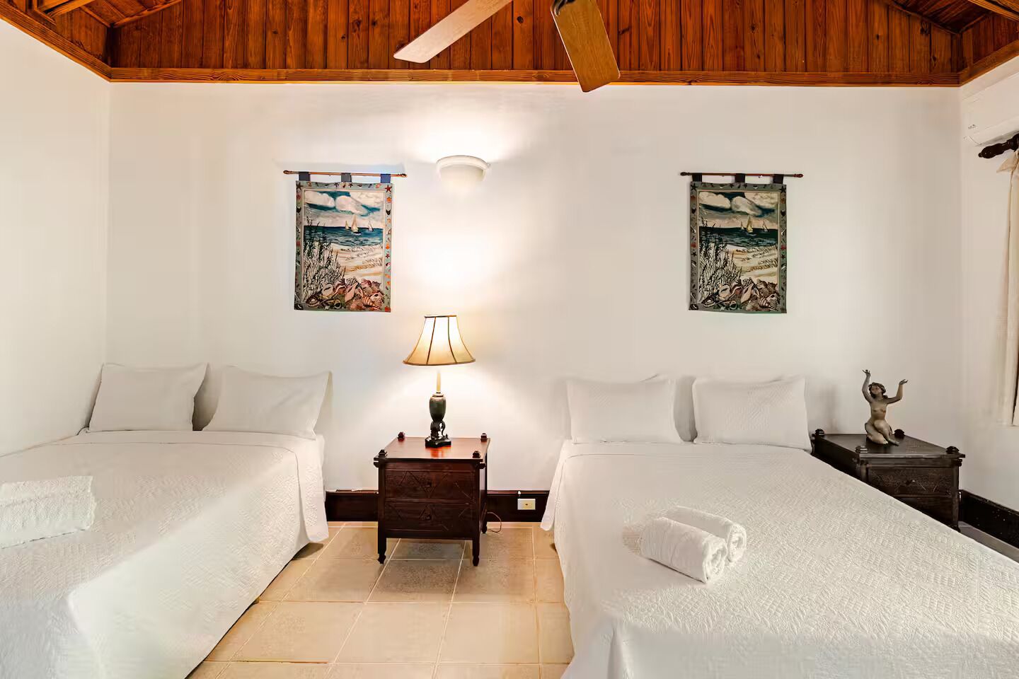 A bedroom with two beds, a nightstand with a lamp, and two paintings on the walls.