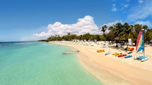 A beach with clear turquoise water, a row of colorful kayaks, and a sailboat.