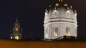 Uma grande cúpula de igreja iluminada com detalhes arquitetônicos elaborados e uma torre de relógio menor ao fundo.