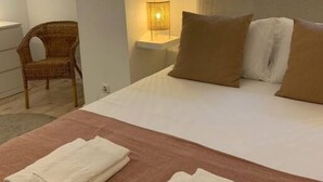 Quarto de hotel com cama, duas almofadas, mesa de cabeceira com abajur, cadeira e janela.