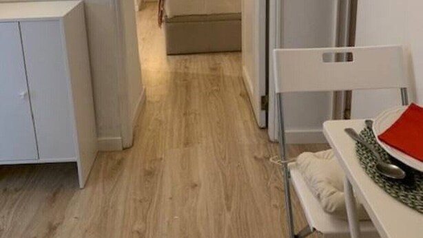 Corredor estreito que dá para um quarto, com cama, mesa de cabeceira e uma cadeira com mesinha.