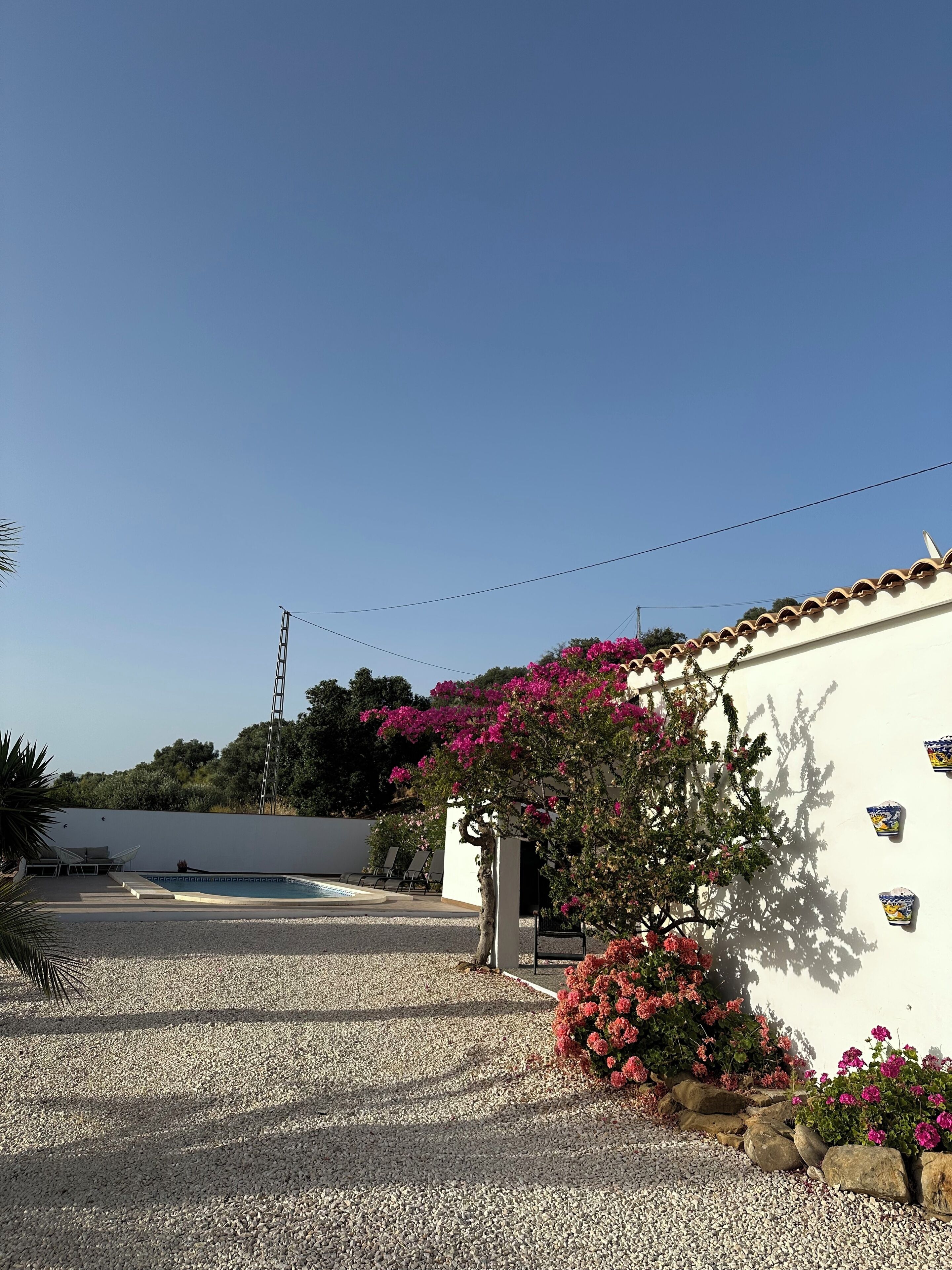 A white building with a tiled roof, a gravel driveway, and a pool area with lounge chairs.