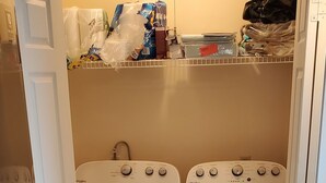 Pesutilassa on kaksi Whirlpool-merkkiä olevaa pesu- ja kuivauskonetta, puulattia ja kuivauskonien yläpuolella oleva hylly, jossa on erilaisia tavaroita.