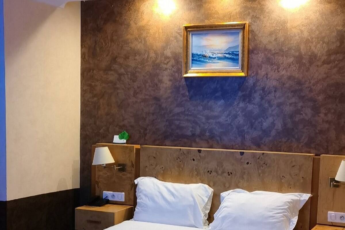 Habitación de hotel con una cama grande, cabecera de madera, dos mesitas de noche con lámparas, un cuadro colgado en la pared y suelo cubierto de alfombra.