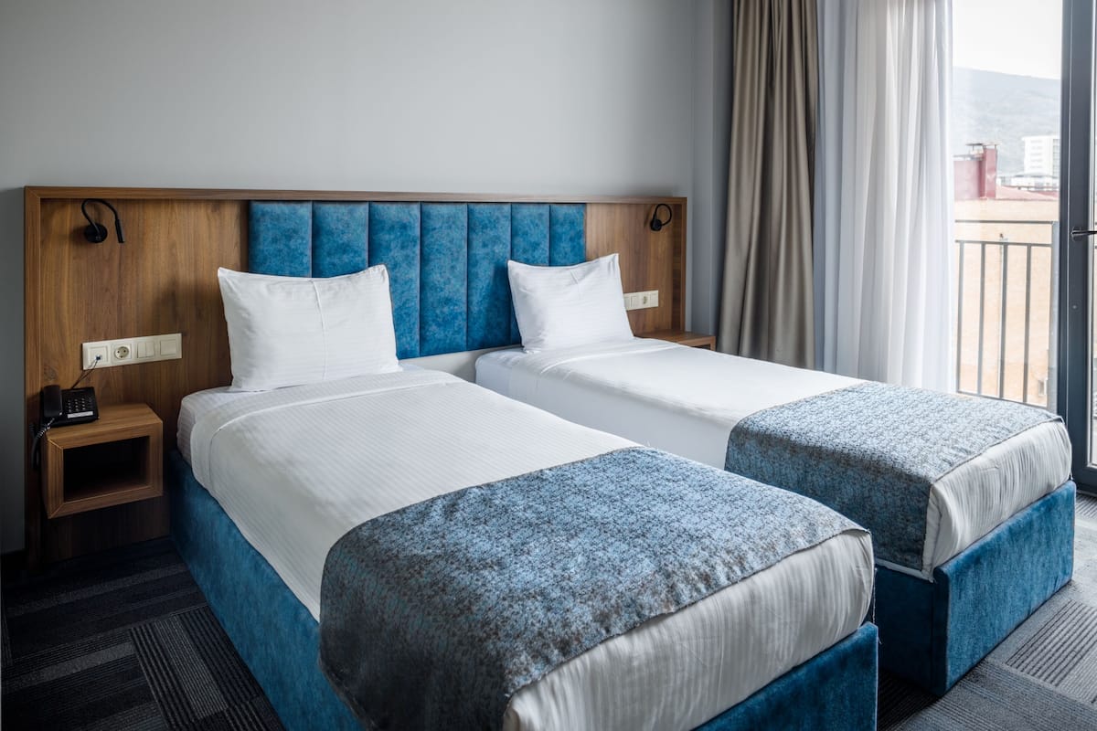 Quarto de hotel com duas camas, cabeceira de madeira, mesa de cabeceira com telefone e vista da cidade através de uma varanda com cortinas.