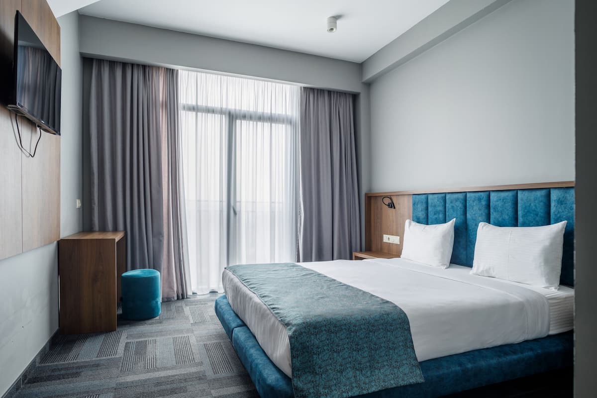 Quarto de hotel moderno com uma cama grande, cabeceira de madeira, mesa de cabeceira pequena e uma janela ampla com cortinas.