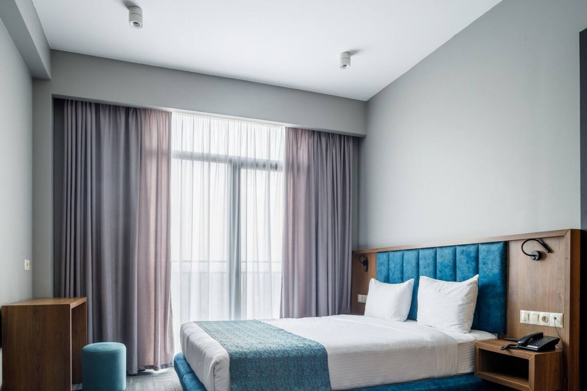 Quarto de hotel moderno com uma cama grande, uma mesinha de cabeceira de madeira, uma escrivaninha e um pequeno banquinho.