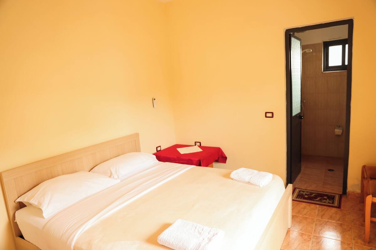 A hotel room with a large bed, a red table, and a wooden door leading to a bathroom.